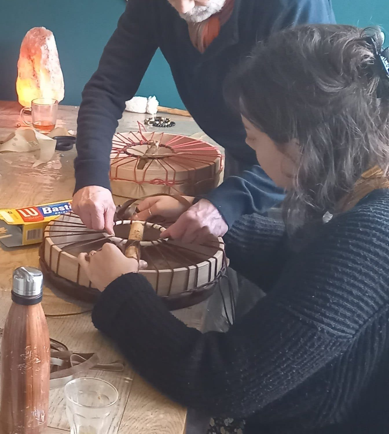 Twee deelnemers spannen samen het touw van een sjamanendrum aan, aan een houten tafel met een zoutlamp erachter